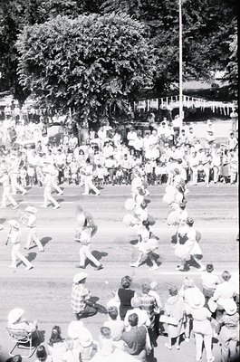 Aerial shot of a lively outdoor festival or parade, likely from the **1960s–1970s**. Crowds gather along a tree-lined street,...