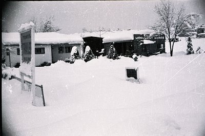 Mid-20th century snowy street scene with light industrial/commercial buildings. Snow-covered signage reads "Toyes" and "Monum...