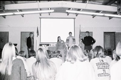 Black-and-white indoor lecture hall scene with four speakers at a podium, facing an attentive audience. Visible text on a T-s...
