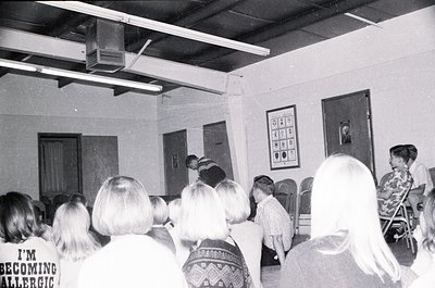 A black-and-white classroom scene from the 1970s–1980s, featuring a group of seated students facing a teacher at a front lect...