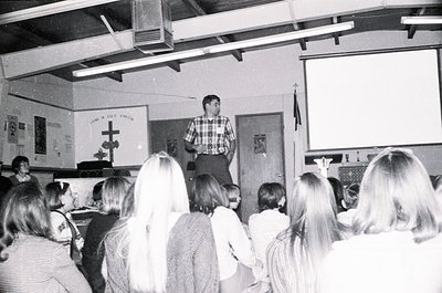A 1970s classroom scene: teacher in plaid shirt addresses seated students. Cross and "Love and God" banner on wall. Industria...