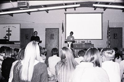 A black-and-white wedding ceremony in a church hall, likely 1970s–1980s. Bride in veil stands at altar with groom beside her;...