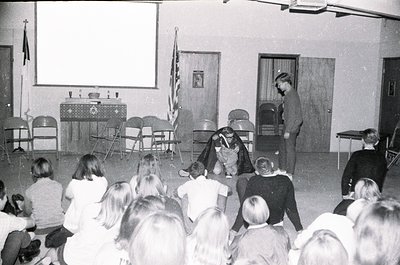 A 1970s-era classroom or assembly hall featuring a puppet show. A performer in costume interacts with a puppet on stage while...