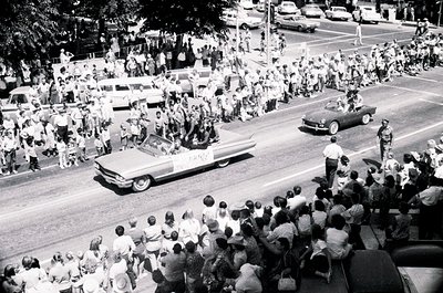 Vintage parade scene featuring a convertible car driving through a dense crowd, likely mid-20th century. Classic cars, specta...