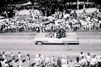 Classic 1960s convertible parade float with "Viva Kennedy" banner, surrounded by dense crowds. Mid-century American street sc...