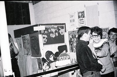 Indoor scene featuring three individuals in a dimly lit, cluttered room covered in posters and political art. Central figure ...