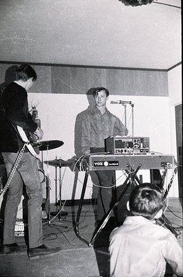Vintage black-and-white photo of a live music performance featuring a guitarist (left) with a Fender-style bass, a keyboardis...