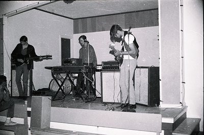Three musicians perform on a modest stage in a dimly lit indoor venue, likely a 1970s rock band. Left: guitarist with acousti...