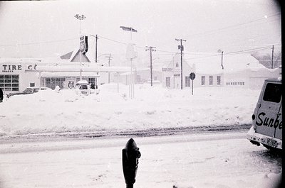 Mid-20th century snowstorm scene: blanketed gas station ("Tire Club") and adjacent buildings under heavy snowfall. Classic 19...