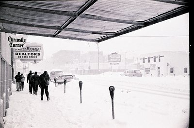 Blizzard conditions at a mid-20th century highway rest stop, featuring a "Curiosity Corner" McDonald’s Realtors Insurance sig...