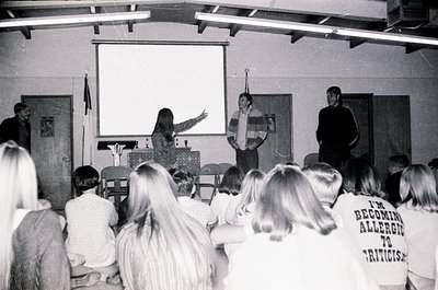 Black-and-white classroom scene featuring three lecturers at a projector screen, addressing seated students. Audience wears 1...