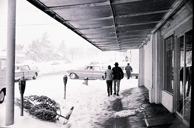 Mid-20th century street scene under a covered sidewalk during heavy snowfall. Three pedestrians in winter coats and hats navi...