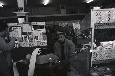 Vintage 1970s-era record store interior with shelves stocked with vinyl albums, posters, and memorabilia. Man in glasses and ...