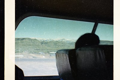 Vintage car windshield view showing snow-capped mountains and a coastal road, likely taken in the 1970s. Distorted lens effec...