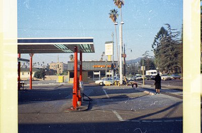 Mid-century gas station with red canopy and white lettering, likely . Palm trees and vintage cars suggest a sunny coastal or ...