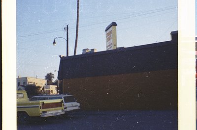 Vintage urban street scene featuring a yellow-and-white station wagon parked beside a dark, flat-roofed building. A sign on t...
