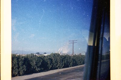 Vintage train window view of rural landscape, featuring dense green shrubbery lining a road. Distant industrial chimney emits...