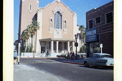 Mid-century Art Deco church corner with vertical stained-glass windows and decorative stonework, flanked by palm trees. "Aval...