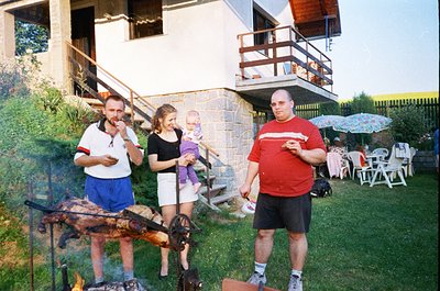Three adults and a child pose outdoors near a stone house with a balcony, grilling skewers over an open fire. The man in red ...