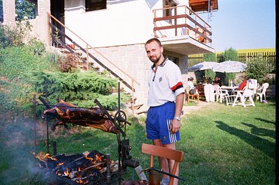 Man in Adidas trackwear grills a whole pig over an open fire in a backyard setting, likely late 20th century. Surrounding are...