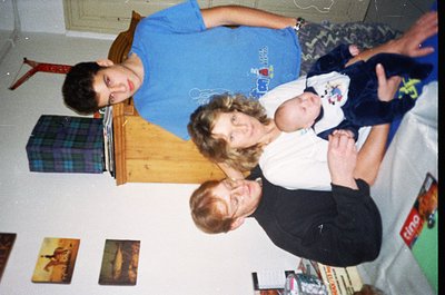 Family portrait in a casual indoor setting, likely late 1990s/early 2000s. Three adults and a baby pose on a bed with a plaid...