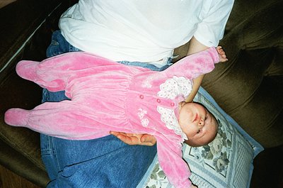 Vintage-style photo of an infant in a pink, ruffled onesie with matching booties, held by an adult’s legs. The setting appear...