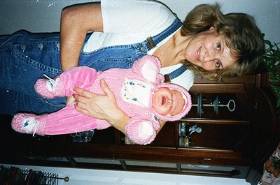 A woman in a denim jacket and blue jeans poses with a baby dressed in a vintage pink knitted outfit, likely from the **1980s–...