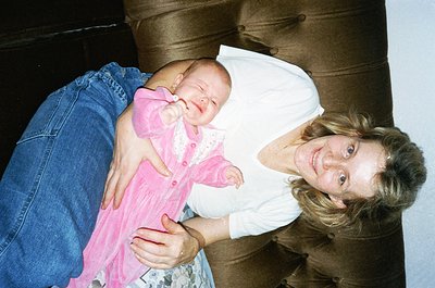 A tender moment captured: a woman cradles a newborn in a pink dress, both smiling at the camera. The scene appears indoors, l...