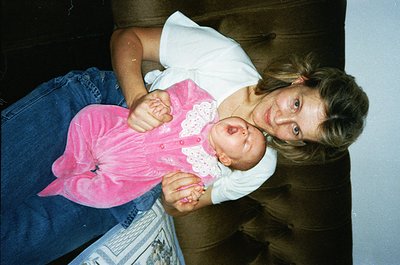 A woman in a white blouse and khaki pants cradles an infant dressed in a vintage pink lace dress, seated on a dark sofa. The ...