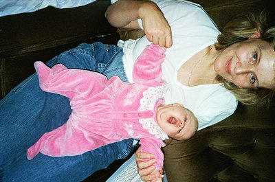 A woman holds an infant dressed in a vintage-style pink lace romper, seated on a wooden bench. The scene suggests a casual, i...