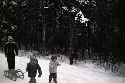 Black-and-white winter scene: three children in winter attire (hooded coats, mittens) playing in a snow-covered forest. One c...