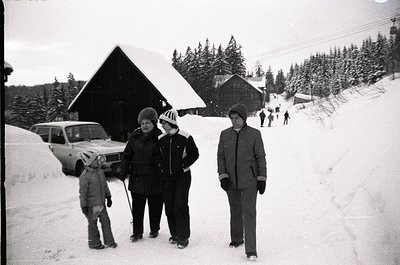 Black-and-white snapshot of a winter outing in a snowy alpine village, mid-20th century. Three adults and a child pose in win...