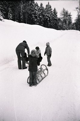 Mid-20th century alpine winter scene: child in vintage sled with adult assisting, others observing. Snow-covered slope framed...