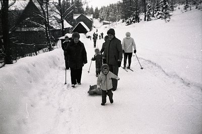 Family skiing on groomed trail in snowy alpine village, mid-20th century. Adults in vintage winter gear (coats, hats) guide c...