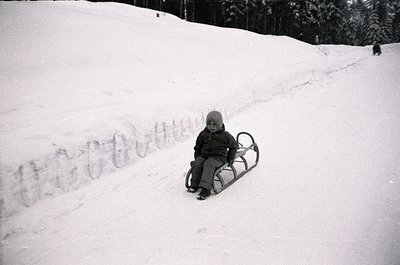 Child in vintage-style sled on snow-covered slope, mid-20th century winter scene. Snow tracks and evergreen trees frame the c...