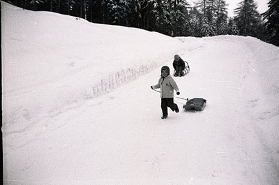 Black-and-white snapshot of two children sledding down a snow-covered hill, likely mid-20th century. Child in foreground push...