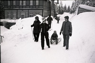 Four adults and a child in winter attire navigate snow-covered alpine terrain, likely a ski resort. Wooden chalet-style build...