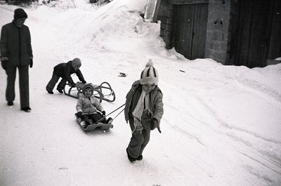 Vintage black-and-white photo of three children sledding on a snow-covered slope beside a wooden structure. Child in foregrou...