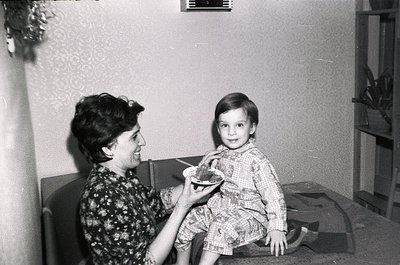 A mid-20th century indoor moment: an adult feeds a young child a small bowl of food in a modestly furnished room. The woman w...