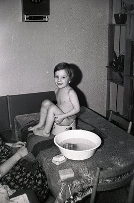 Young child sitting on a patterned sofa in mid-20th century indoor setting, holding a white enamel basin. Soap and bar of soa...