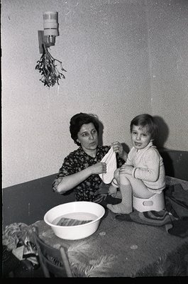 A black-and-white photo capturing a mid-20th-century domestic moment: a woman in patterned dress assists a toddler in a train...