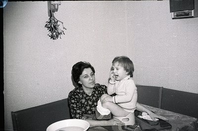 Vintage black-and-white photo of a woman cradling a toddler in a modest indoor setting, likely mid-20th century. Patterned fl...