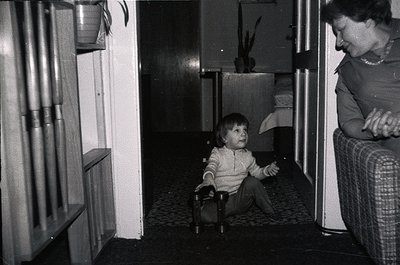 A mid-20th century indoor scene captures a child seated on a highchair in a dimly lit hallway, flanked by a woman in patterne...