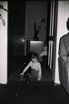 Black-and-white interior shot of a child playing with a small object on tiled floor, framed by white-painted walls and doors....