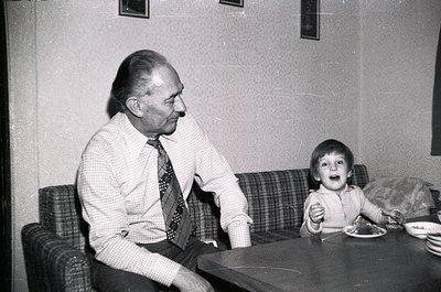 Mid-20th century indoor portrait: elderly man in checkered shirt and patterned tie sits beside a young boy at a wooden table....