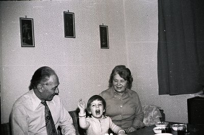 Black-and-white indoor portrait of a family: elderly man in suit, woman in patterned blouse, and child laughing mid-table. Fr...