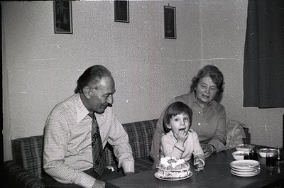 Vintage black-and-white indoor family moment: elderly man in checkered shirt and tie, child in white dress eating birthday ca...