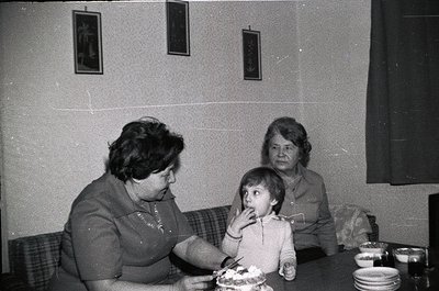 Two women and a young child seated indoors at a table, likely mid-20th century. The woman on the left feeds the child a slice...