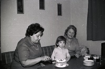 Black-and-white indoor scene featuring three generations: two women (likely mother and grandmother) and a child at a round ta...
