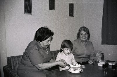 Two women and a child share a cake-cutting moment in a mid-century interior. The woman on the left wears a dark blouse with a...